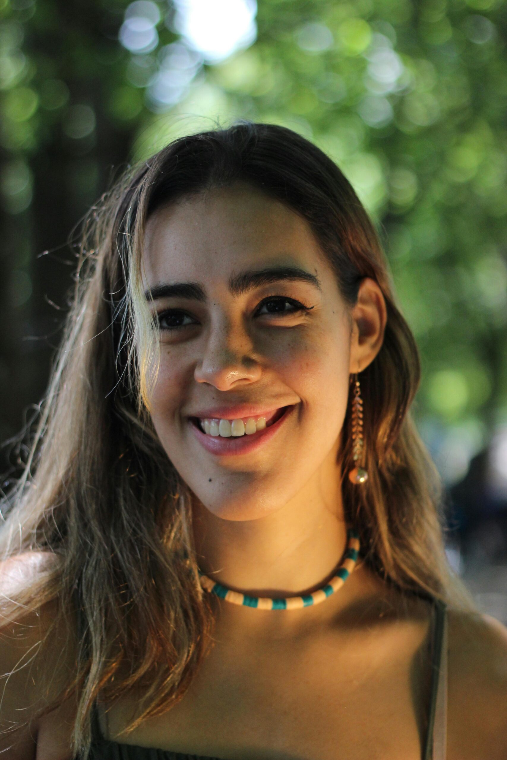 Portrait of a woman smiling warmly in a sunny outdoor setting, surrounded by greenery.